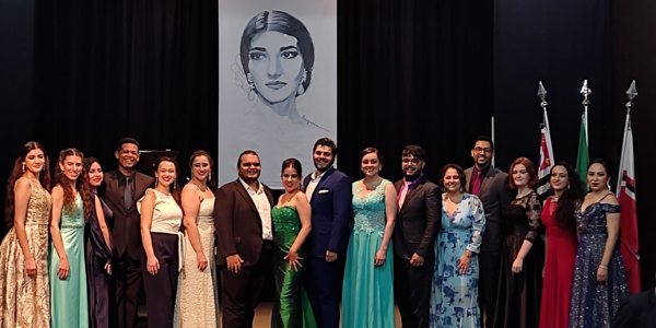 Palco do Concurso de Canto Maria Callas. Os 16 cantores premiados estão enfileirados, vestindo roupas formais e sorrindo para a foto.